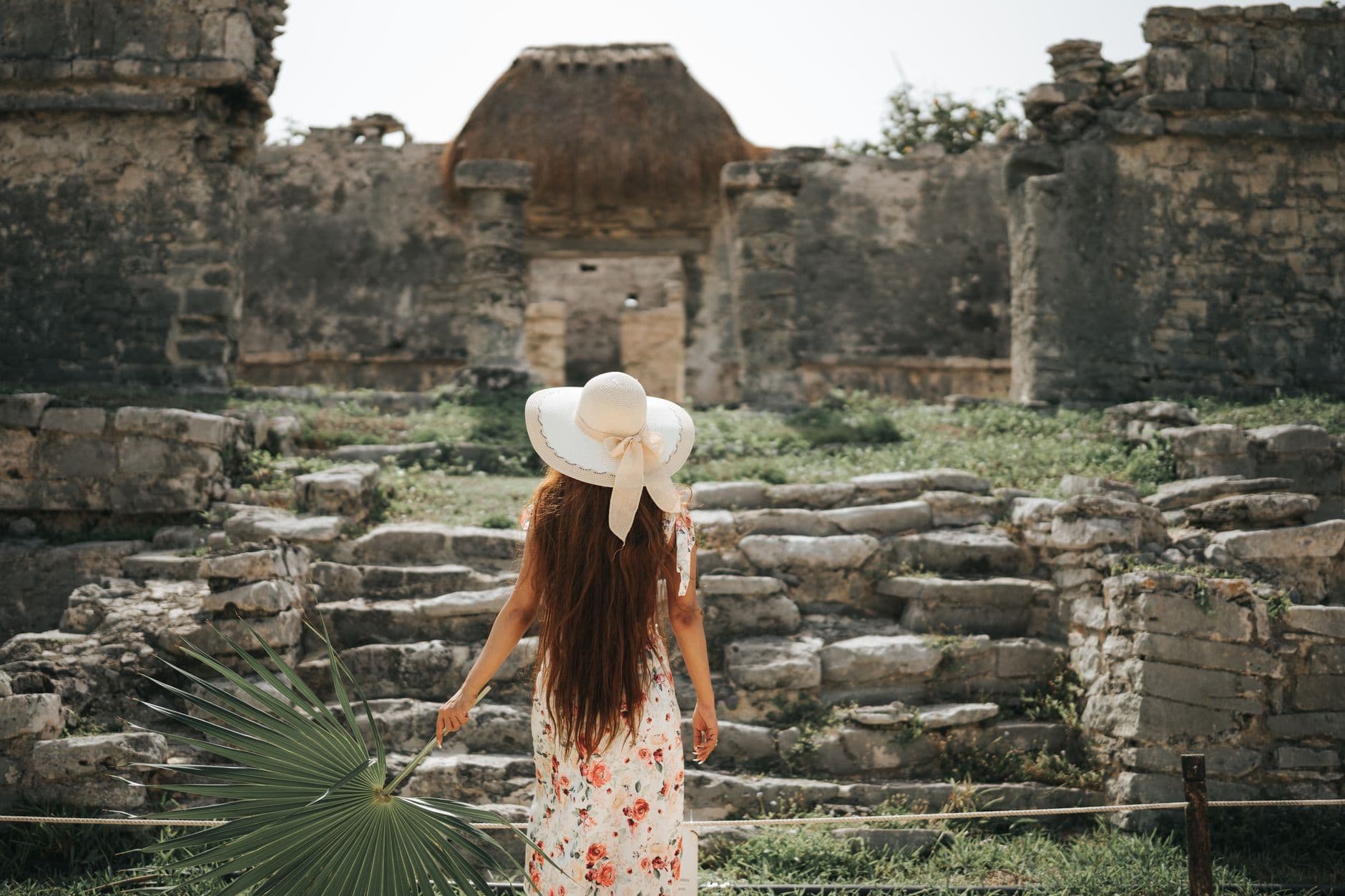A woman in a floral dress and sun hat explores ancient stone ruins on a sunny day.